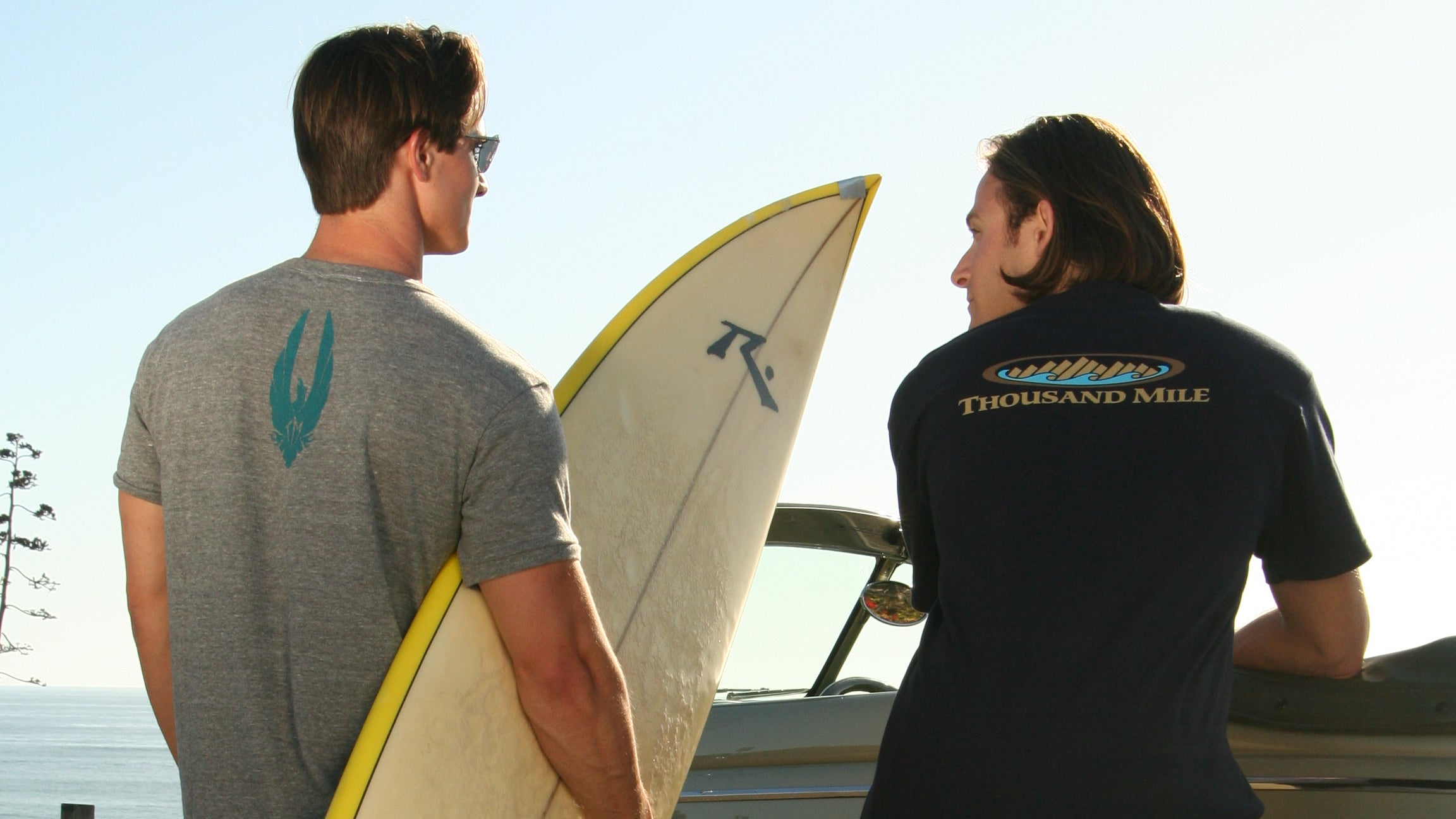 Two men standing outdoors beside a vintage car, one holding a surfboard. One man wears a gray t-shirt with a teal graphic and black shorts, while the other wears a dark navy t-shirt with a colorful graphic and white shorts.